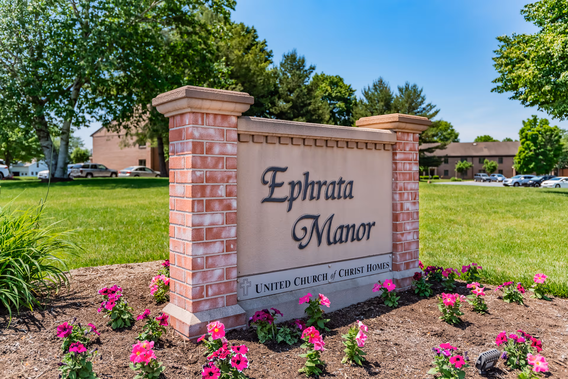 Brick and stone sign reading "Ephrata Manor" surrounded by flowers with the facility buildings and lawn in the background.