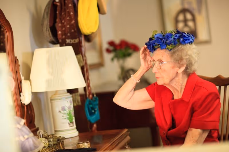 An elderly woman wearing glasses and a red blouse is sitting at a wooden vanity table, adjusting a blue flower crown on her head. The room has a lamp, a mirror, and various personal items on the table, with a blurred background featuring a coat rack and a vase of red flowers.