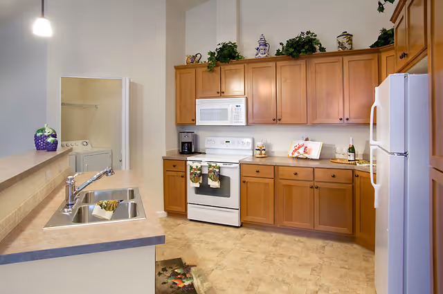 A clean and well-lit kitchen with wooden cabinets, a white stove with a microwave above it, a white refrigerator, a double sink with a chrome faucet, and a countertop with decorative items. In the background, there is a small laundry area with a washer and dryer visible through an open door.