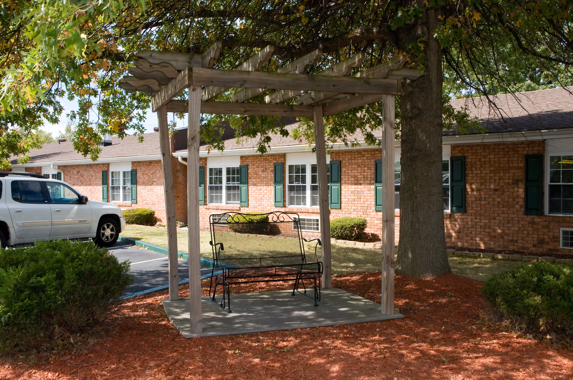 A wooden pergola with a metal bench swing under a tree in front of a single-story brick building with green shutters and a parked white SUV.