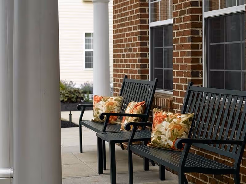 Outdoor seating area with black metal benches adorned with floral patterned cushions, situated on a concrete patio next to a brick building with large windows and white columns.