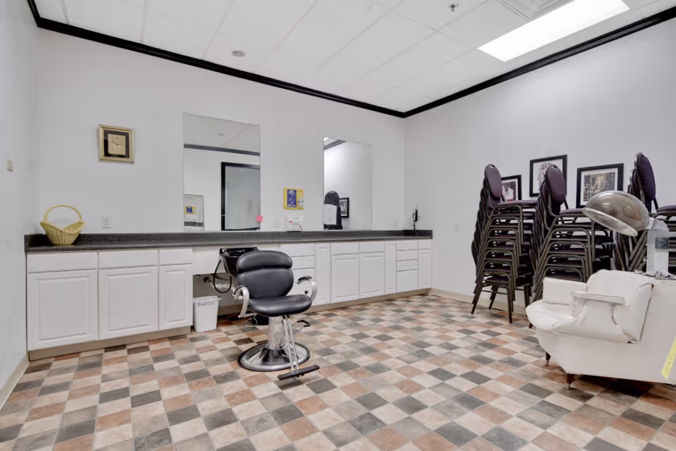 A salon-style room with a barber chair in front of mirrors, long white cabinetry, stacked chairs, and a patterned tile floor.