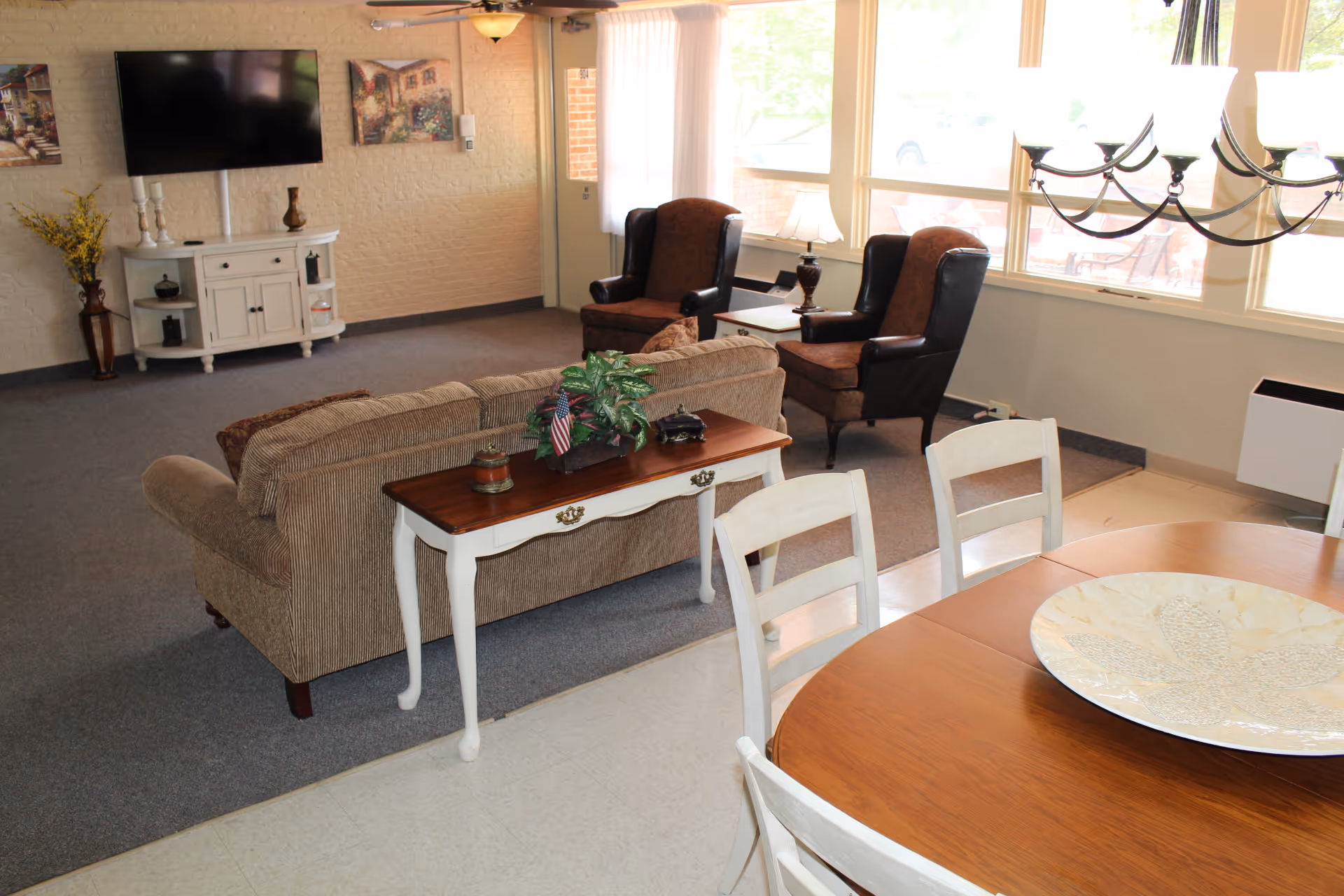 A cozy living room area with a beige sofa, two brown armchairs, a white console table with decorative items, a wall-mounted flat-screen TV, and a dining table with white chairs and a decorative plate. Large windows let in natural light.