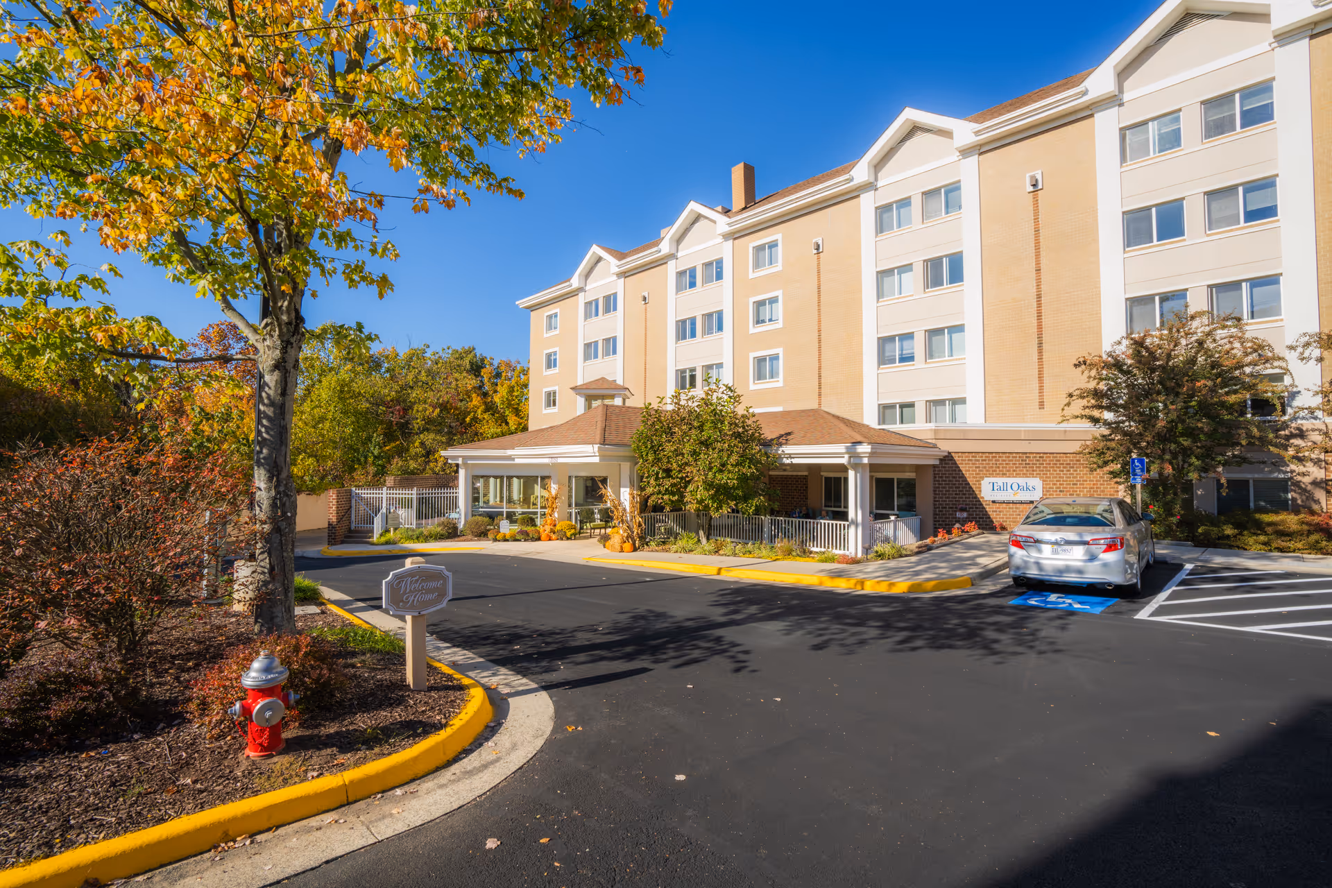 Front entrance of the Tall Oaks Assisted Living building with a driveway, parked car, and autumn trees.