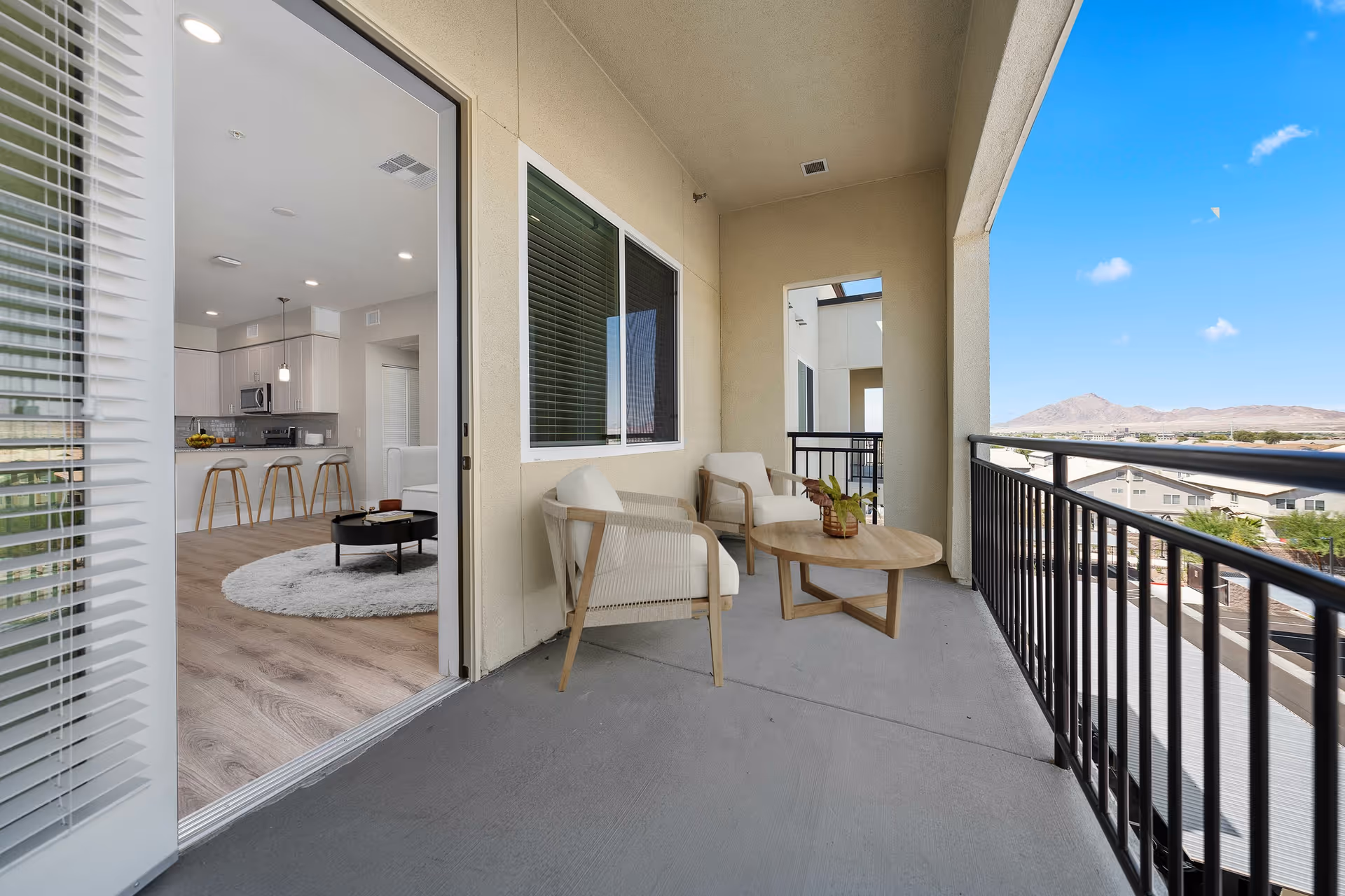 Balcony area with two cushioned chairs and a round wooden table with a plant on it. The balcony has a black metal railing and overlooks a neighborhood with mountains in the distance under a blue sky. Through the open sliding glass door, a modern kitchen and living room with bar stools, a round rug, and a coffee table are visible.
