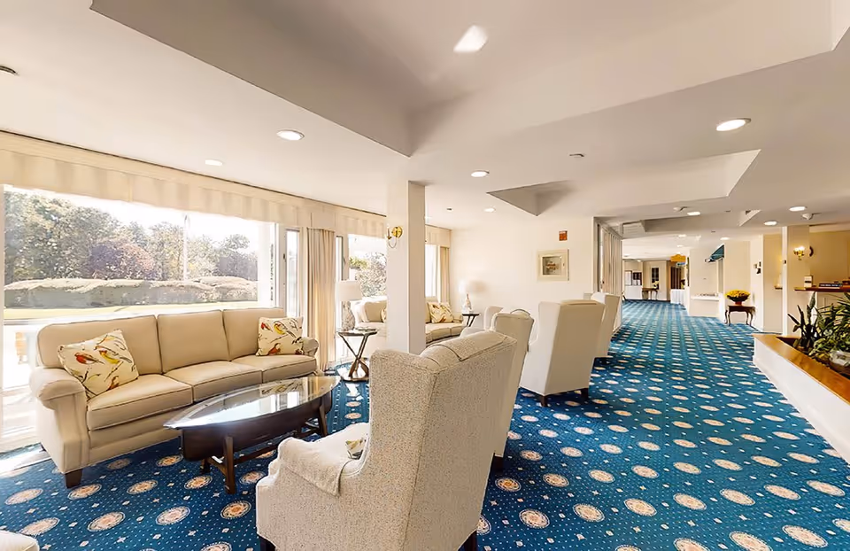 A bright and spacious living room area in Thirwood Place featuring beige sofas and armchairs arranged around a glass coffee table. Large windows allow natural light to fill the room, highlighting the blue patterned carpet and white walls. The space extends into a hallway with additional seating and decorative plants.