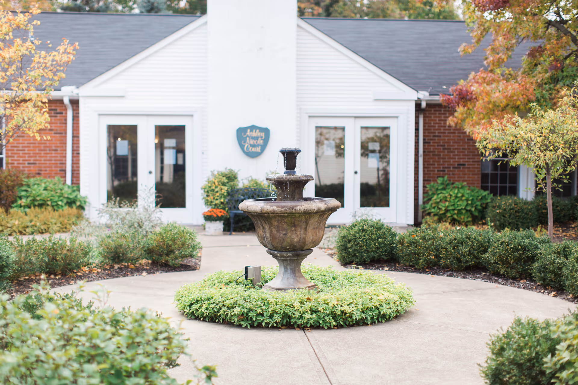 A stone tiered fountain sits in the center of a circular walkway surrounded by shrubs in front of the white entrance of a brick building.