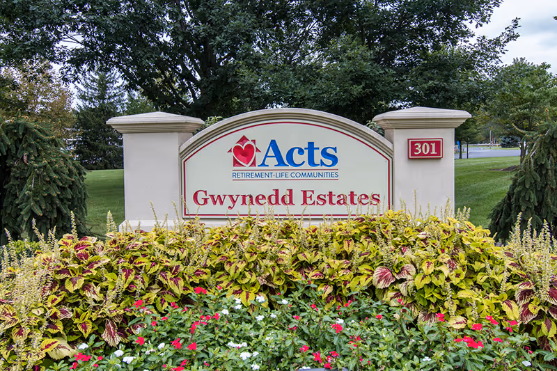 Outdoor sign for Acts Retirement-Life Communities, Gwynedd Estates, surrounded by green shrubs and colorful flowers with trees and grass in the background.