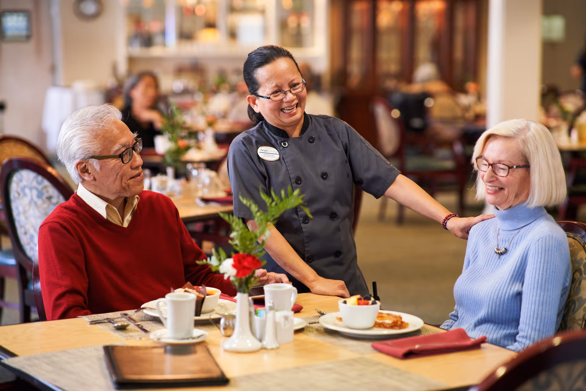A smiling staff member in a dark uniform interacts warmly with two elderly residents seated at a dining table set with plates, cups, and a flower vase in a well-lit dining room.