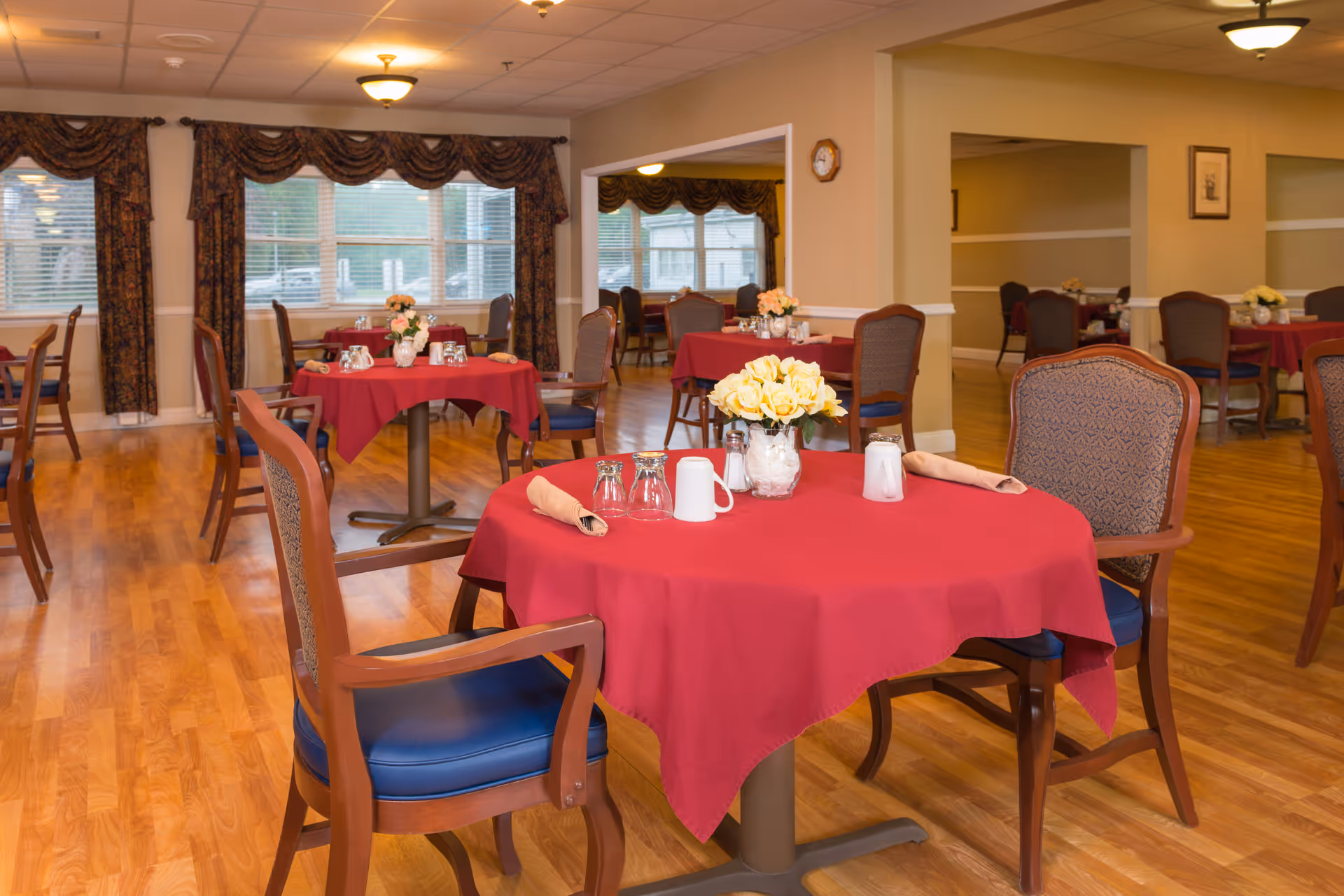 A dining room with multiple round tables covered with red tablecloths, each set with napkins, glasses, and flower centerpieces. The room has wooden floors, large windows with patterned curtains, and several chairs with wooden frames and blue cushions.