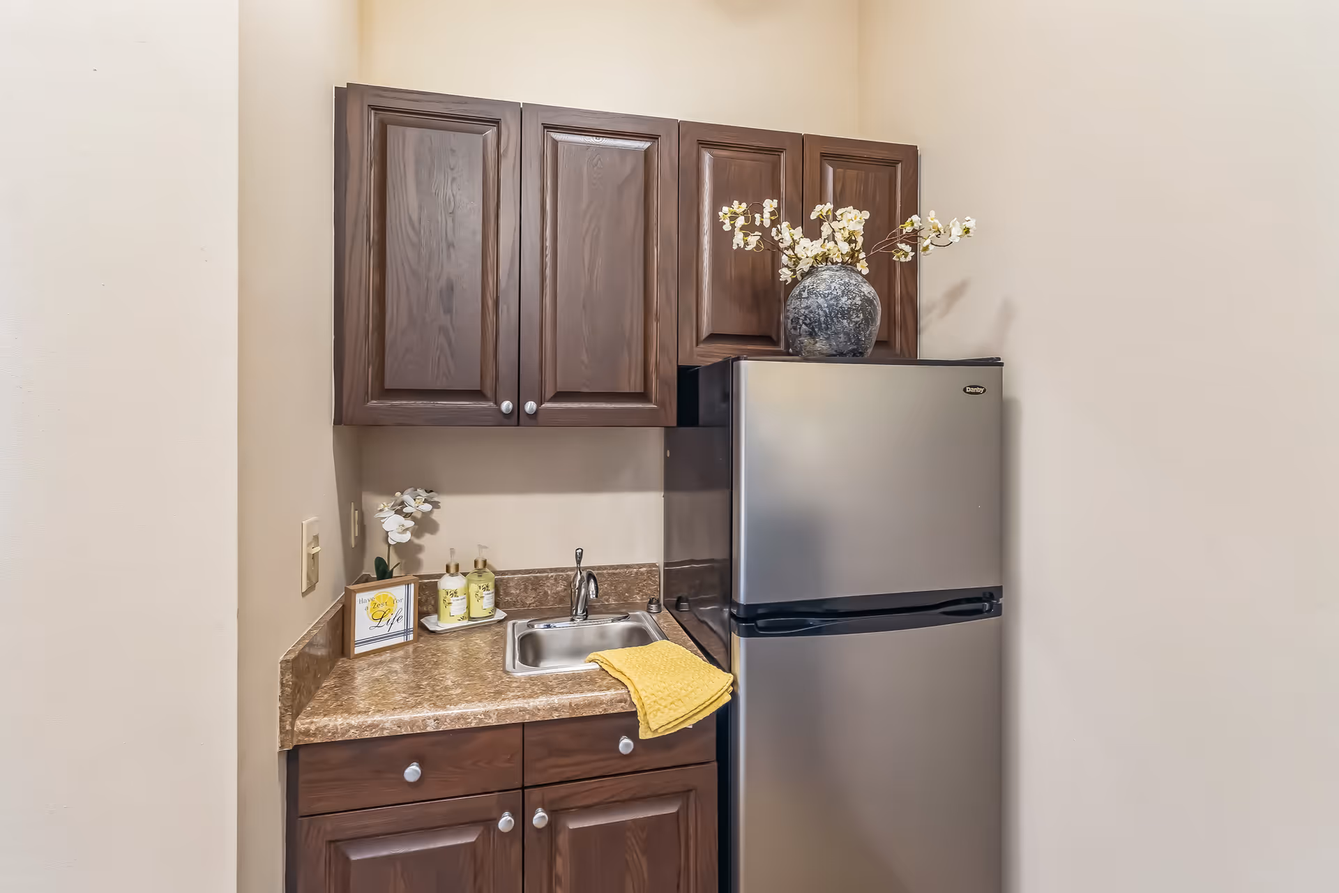 Small kitchenette area with dark wooden cabinets, a stainless steel mini refrigerator, a small sink with a yellow towel draped over the edge, a decorative vase with white flowers on top of the refrigerator, and two bottles of soap or lotion on the countertop next to a small framed sign.