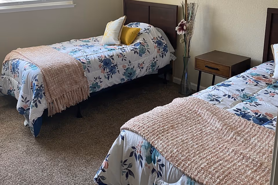 Two twin beds with floral bedding and pink throws flank a wooden nightstand and a tall vase in a carpeted bedroom.