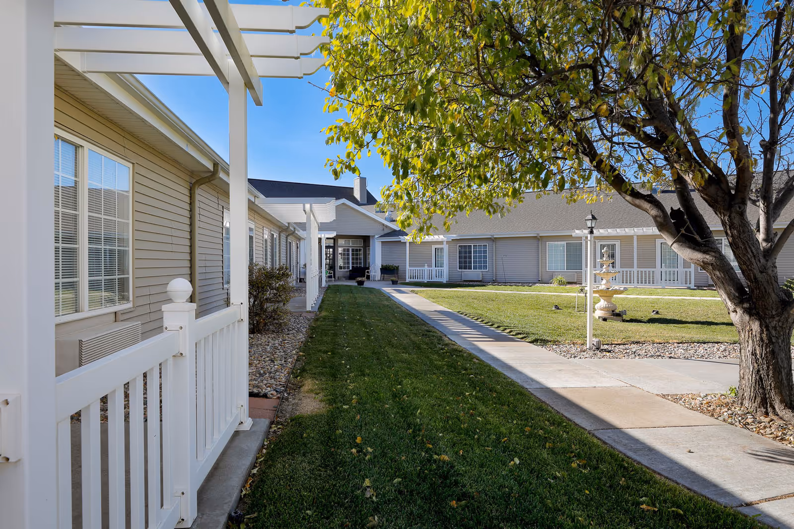 Outdoor courtyard area of Homestead Assisted Living of Hays featuring a green lawn, a tree, a concrete walkway, beige siding buildings with white trim, and a decorative fountain.