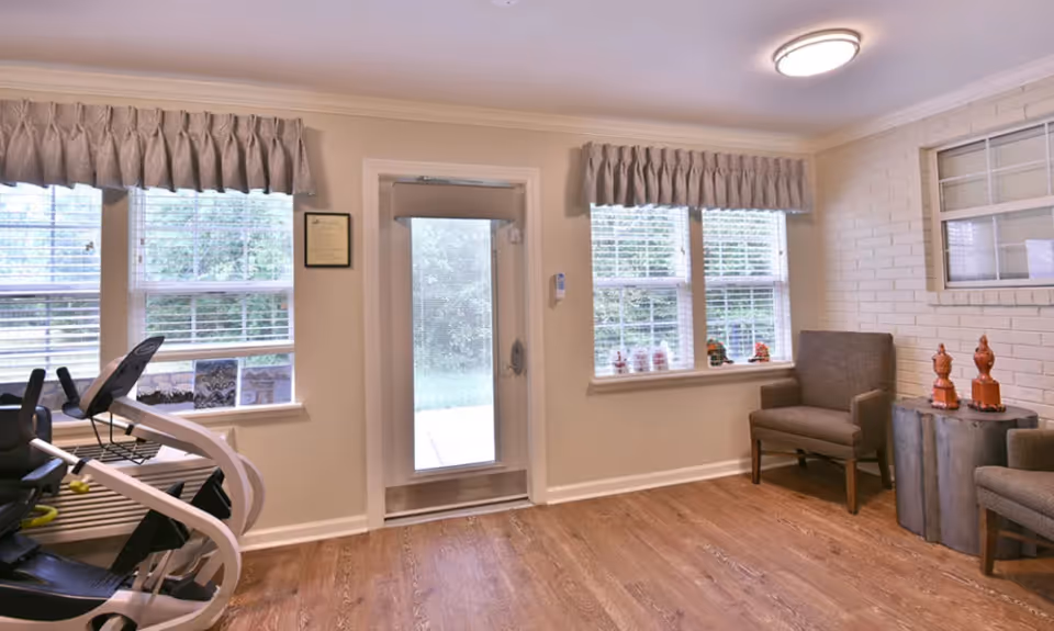 A bright room with wooden flooring featuring two gray armchairs and a small round table with decorative items. There are two windows with blinds and valances, and a glass door leading outside. Part of an exercise machine is visible on the left side.