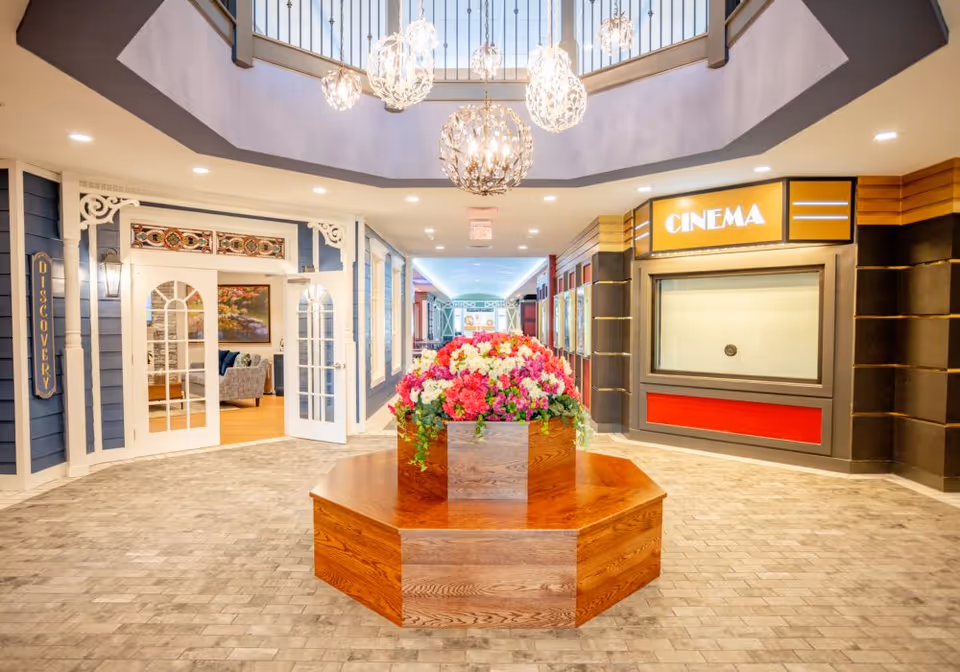 Interior view of a senior living facility hallway with a central wooden planter filled with colorful flowers. To the left, there is an entrance labeled 'Discovery' with white double doors and decorative trim. To the right, there is a cinema entrance with a bright yellow sign. Above, several modern spherical chandeliers hang from a skylight ceiling.