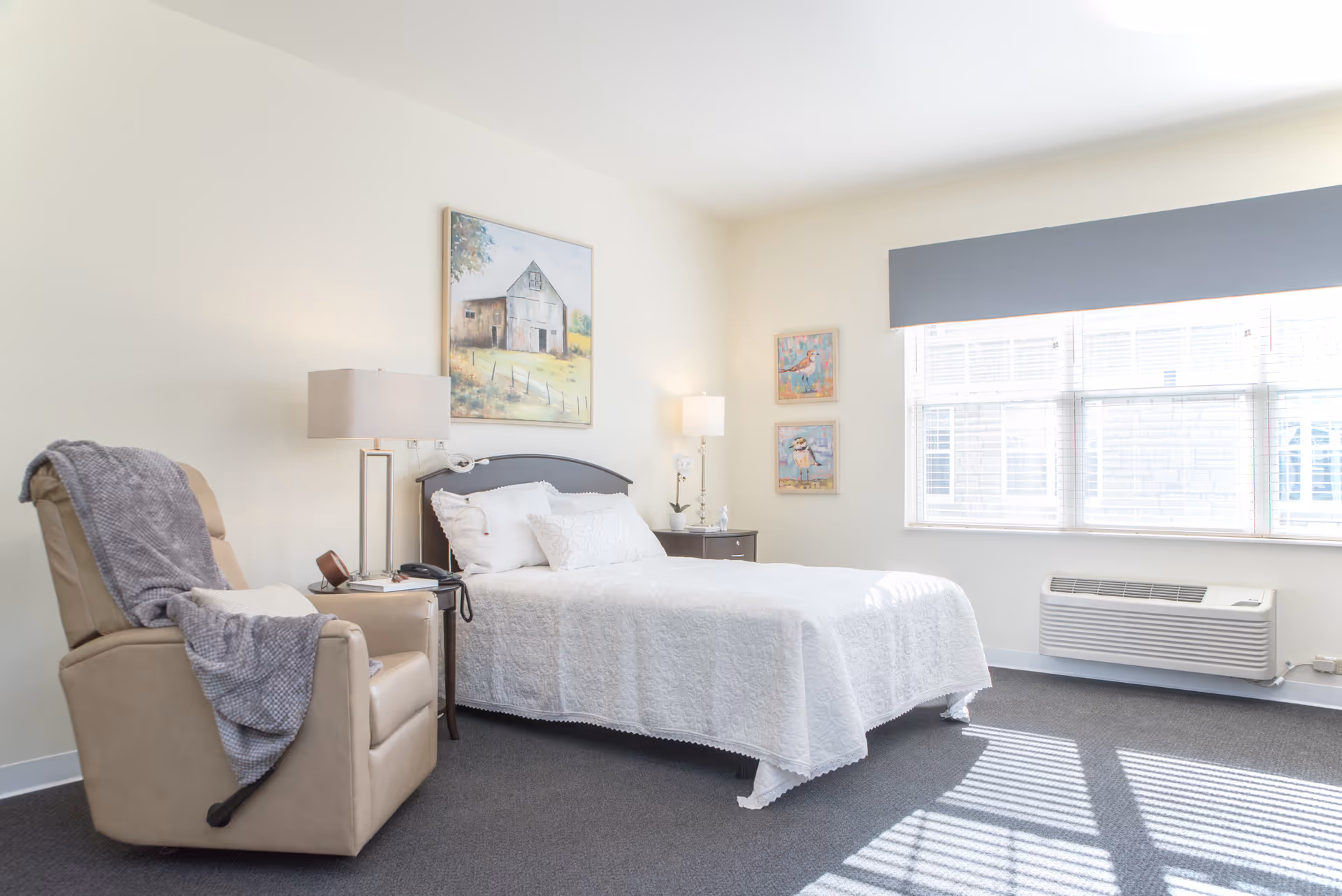 A bright and clean bedroom with a bed covered in a white quilt, a beige recliner chair with a gray blanket draped over it, two bedside tables with lamps, and three framed pictures on the walls. A large window with blinds allows natural light to fill the room.