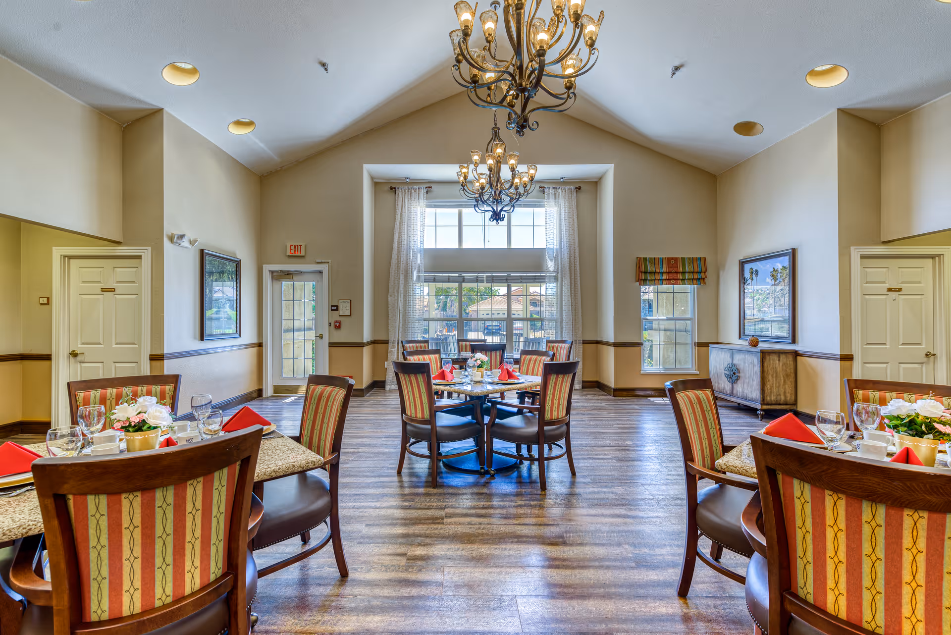 Bright dining room with multiple set tables and striped chairs, chandeliers, and a large window at the far end.