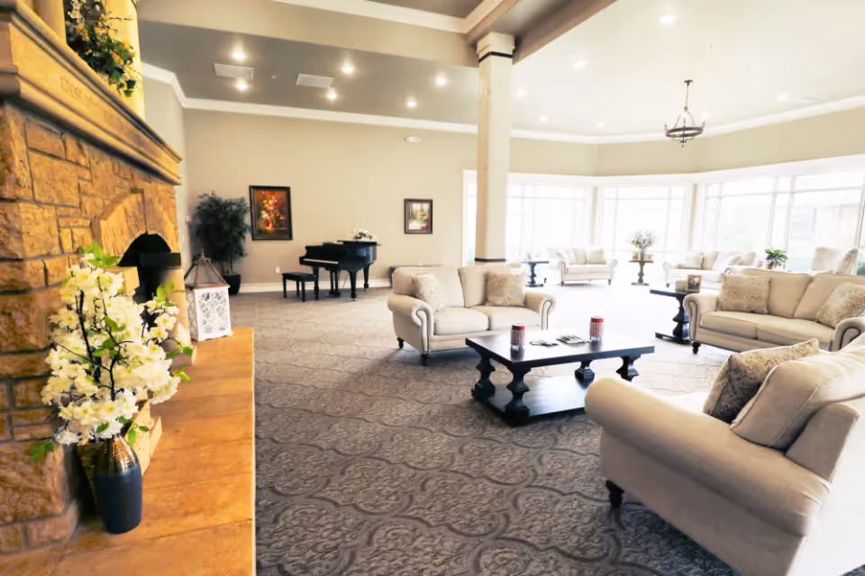 Spacious and well-lit living room area with beige sofas arranged around a dark wooden coffee table, a stone fireplace adorned with white flowers on the left, a grand piano in the background, and large windows letting in natural light.