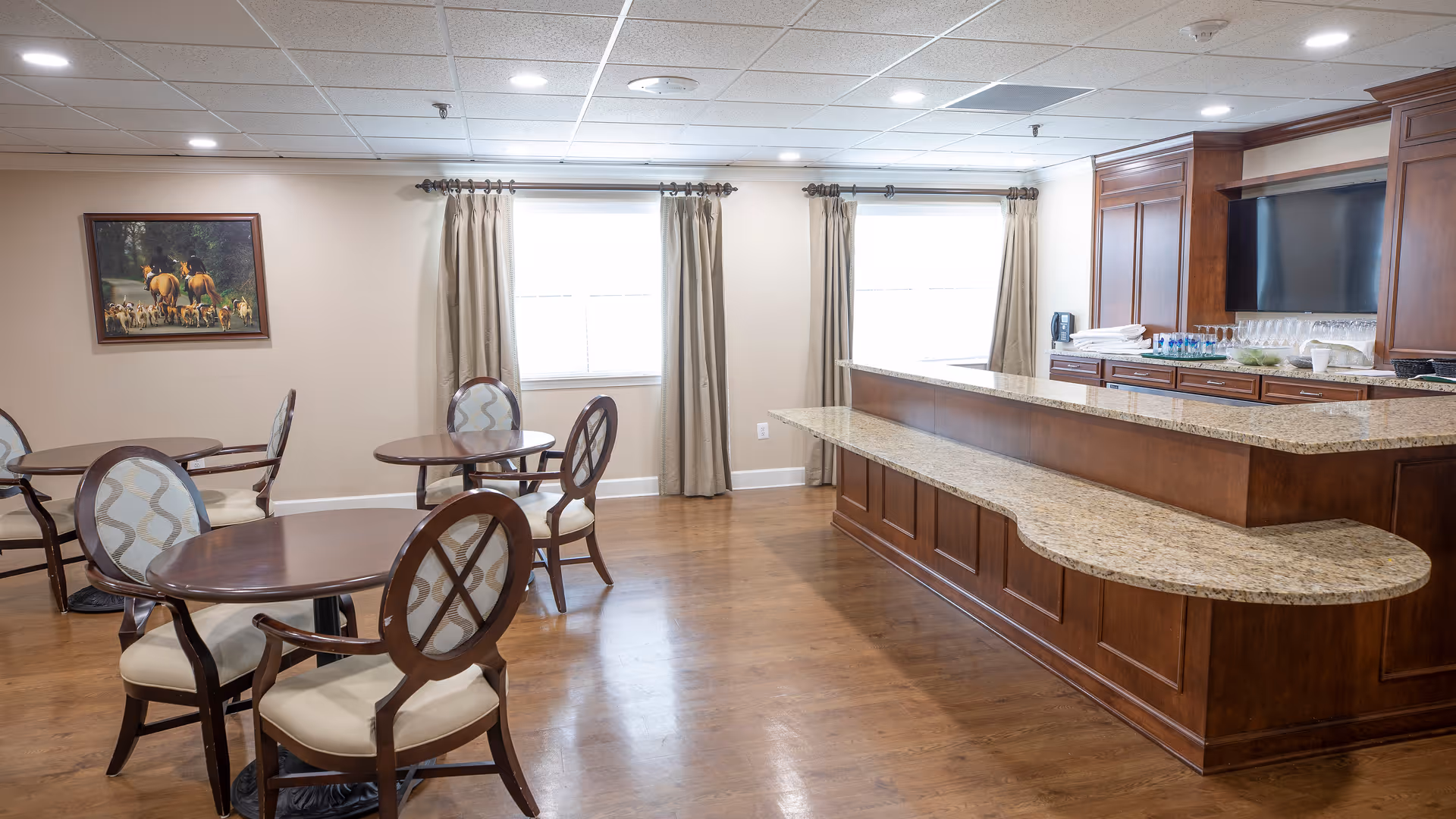 A bright and clean common dining area with wooden floors, several round tables with cushioned chairs, a large wooden counter with granite countertop, and a wall-mounted TV. Two windows with beige curtains allow natural light into the room. A painting depicting people on horseback with dogs hangs on the wall.