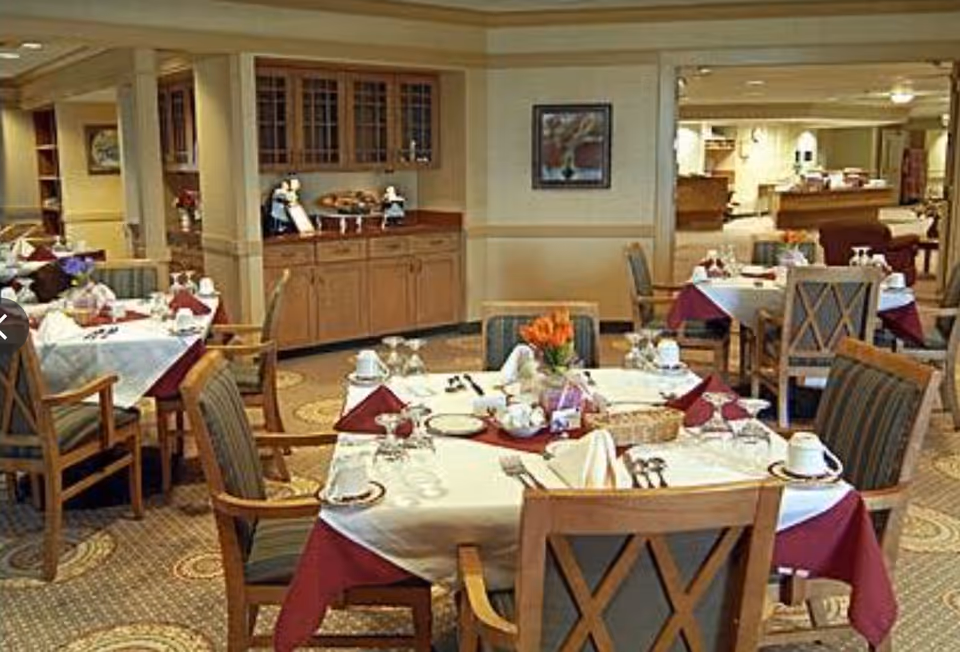 A dining room in a senior living facility with tables set for a meal. Each table is covered with a white tablecloth and a burgundy overlay, and is set with plates, cups, silverware, and folded napkins. There are wooden chairs with cushioned seats around the tables. In the background, there is a wooden cabinet with glass doors and a countertop displaying decorative items. The room has carpeted floors with a circular pattern and soft lighting.