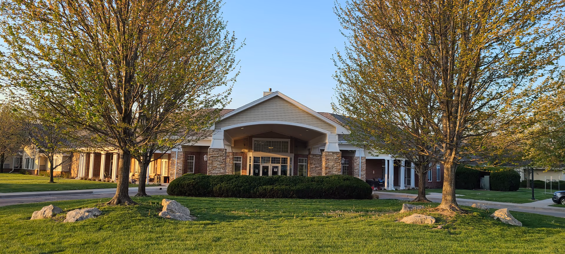 Front exterior view of a single-story senior living facility building named Villa Maria, with a covered entrance supported by stone pillars, surrounded by green lawns, trees with fresh leaves, and rocks on the grass under a clear blue sky.