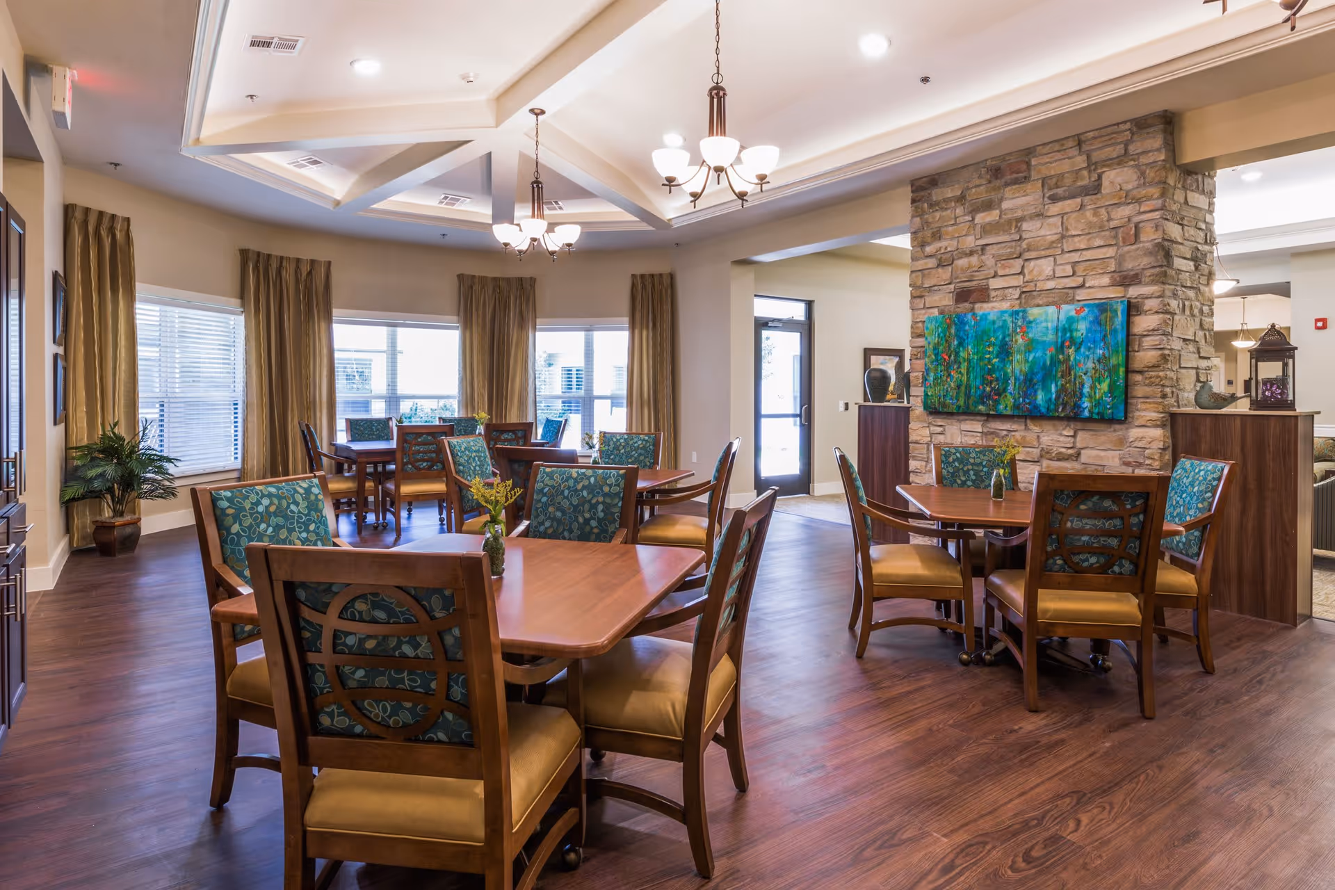 A bright and spacious dining room in an assisted living facility with wooden tables and chairs featuring blue patterned cushions. The room has large windows with beige curtains, a stone accent wall with colorful artwork, and elegant ceiling lights. The floor is a dark wood laminate, and there are decorative plants and vases on the tables.
