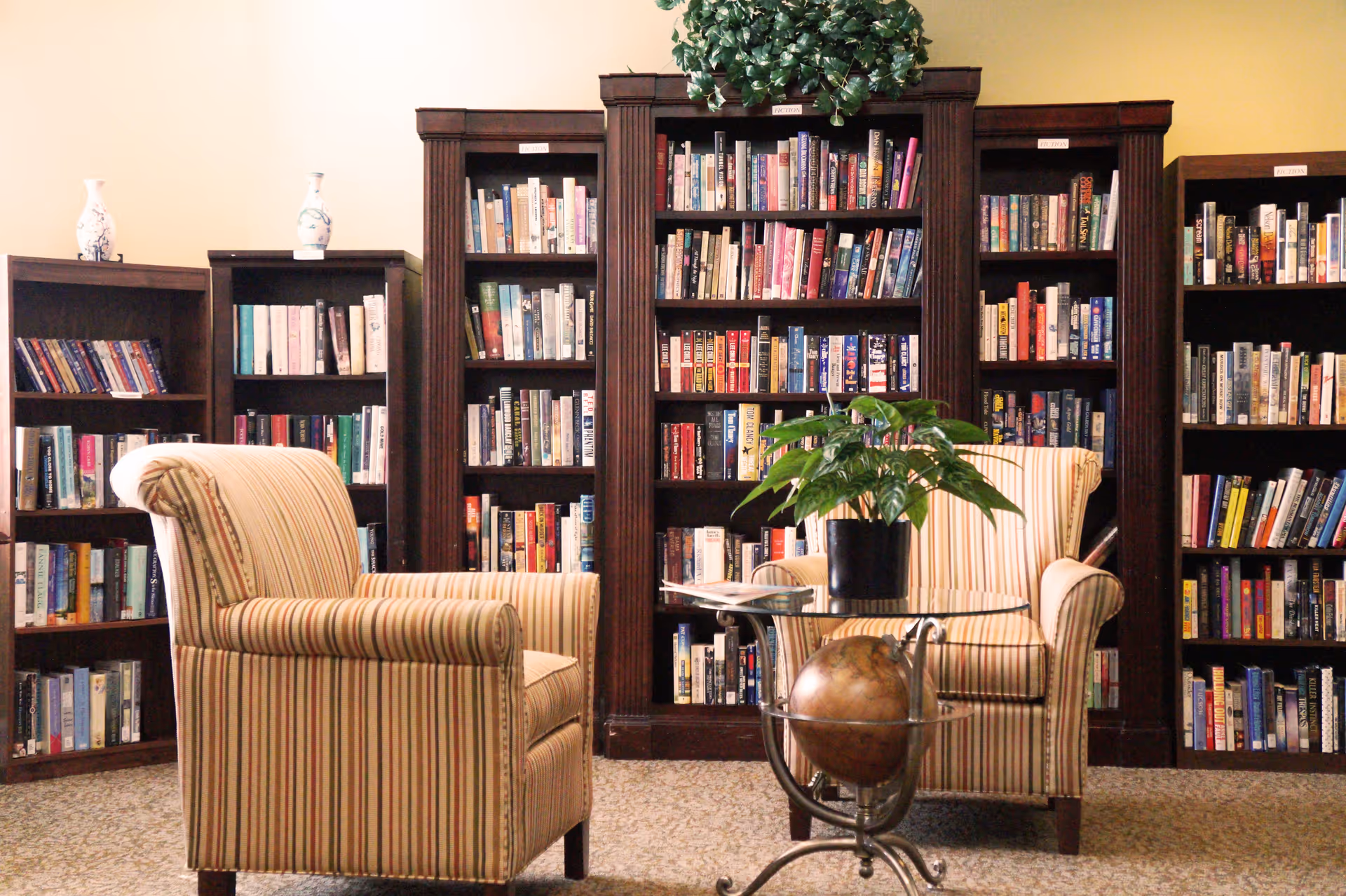 A cozy reading area with two striped armchairs facing each other, a glass-top table with a globe underneath and a potted plant on top, and several dark wood bookshelves filled with books in the background.