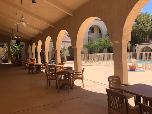 Outdoor covered patio area with multiple wooden tables and chairs under a ceiling with fans, adjacent to a fenced swimming pool and surrounded by beige stucco buildings with arches and greenery.