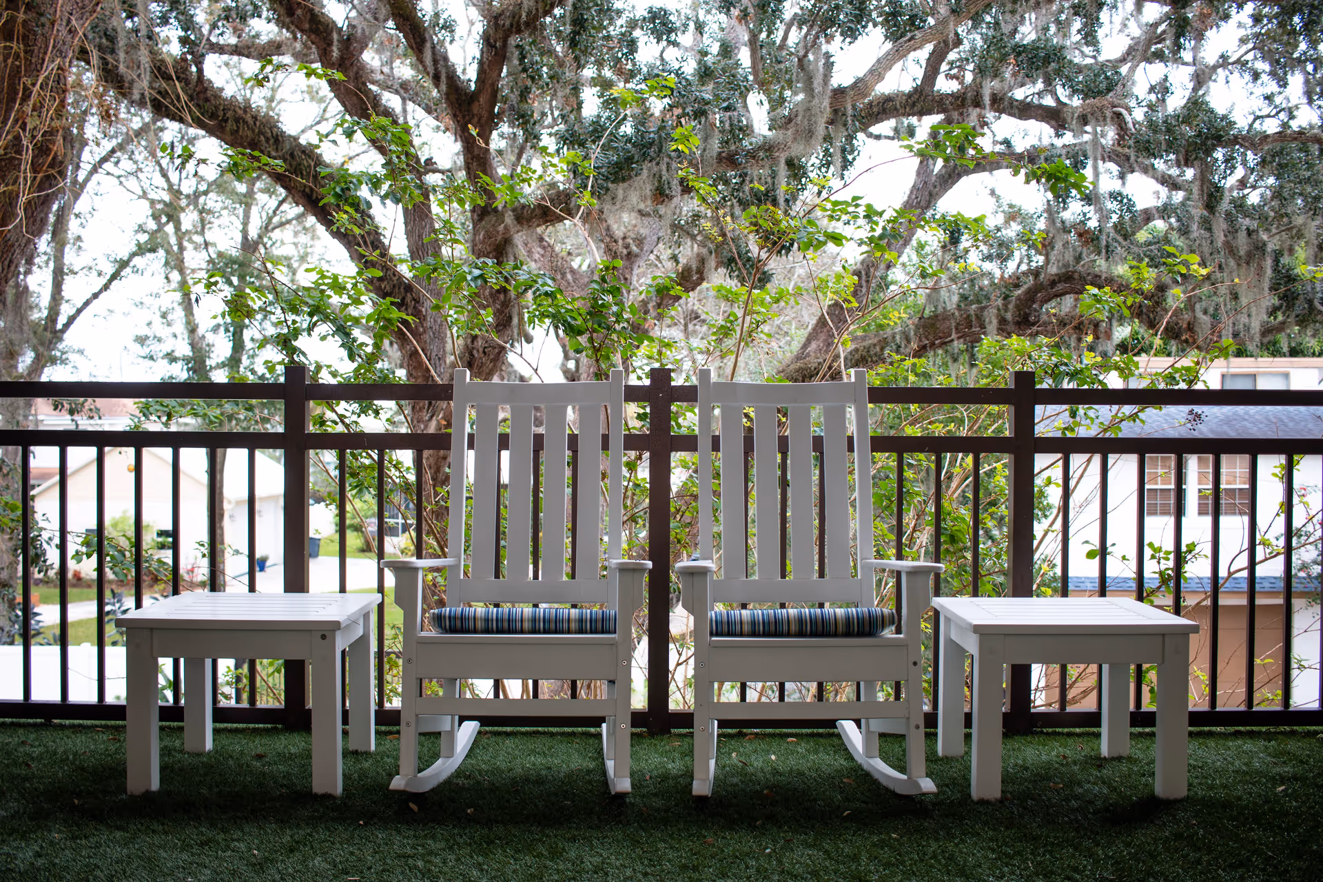 Two white wooden rocking chairs with striped cushions on a green artificial grass floor, flanked by two small white tables, on a balcony with a black railing and large trees with hanging moss in the background.