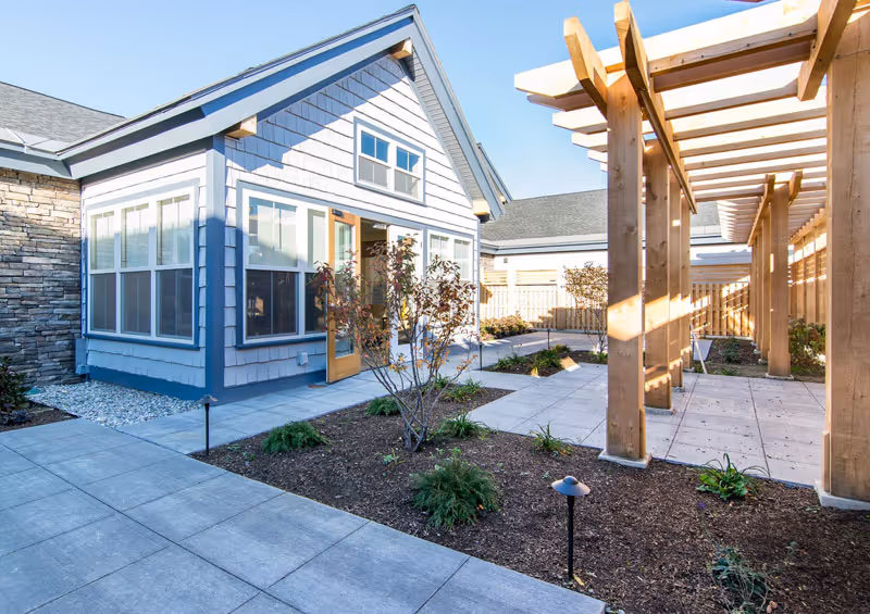 Outdoor courtyard area at Maple Ridge Memory Care featuring a paved walkway, a wooden pergola, small landscaped garden beds with plants, and a building with large windows and a door opening to the courtyard.