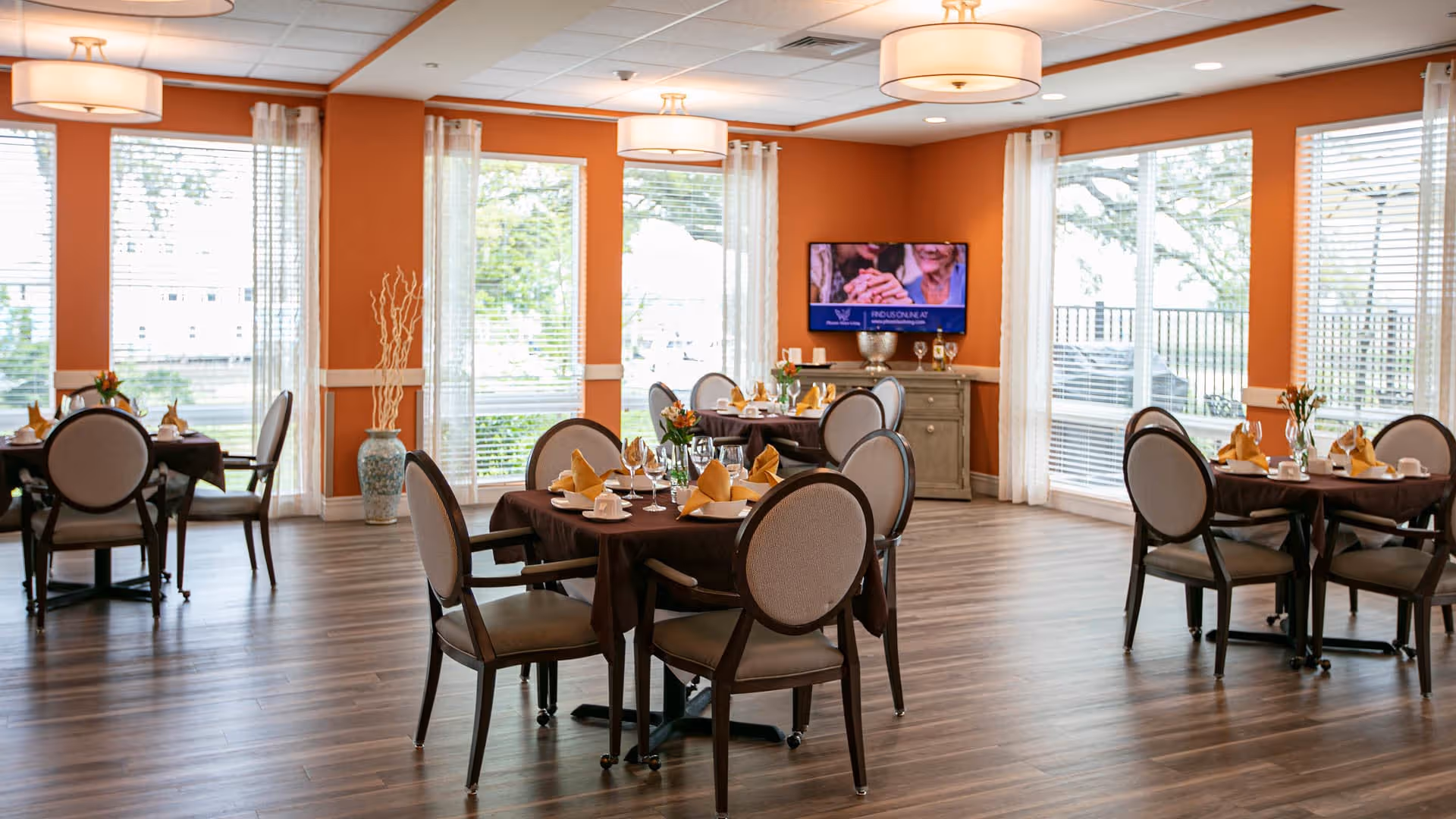 A bright dining room with several round tables covered with dark tablecloths, each set with plates, glasses, napkins, and small flower arrangements. The room has large windows with white curtains, orange walls, and wood flooring. A TV is mounted on the far wall above a small cabinet.