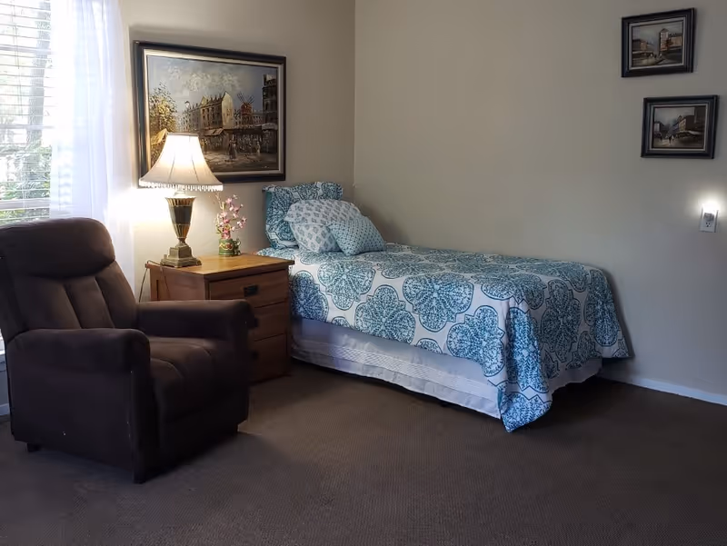 A cozy bedroom with a single bed covered in a blue and white patterned bedspread and matching pillows. Next to the bed is a wooden nightstand with a lamp and a small flower vase. A comfortable dark brown armchair is positioned near a window with sheer white curtains. The walls are decorated with framed paintings.