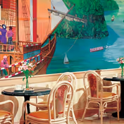 Indoor seating area with round tables and wicker chairs featuring orange cushions. Each table has a small vase with flowers. The background shows a large colorful mural depicting a sailing ship on water with green hills and a small boat.
