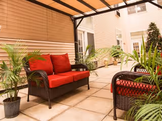 Outdoor patio area with wicker furniture featuring red cushions, surrounded by potted green plants and covered by a pergola. The background shows the exterior walls and windows of the facility.