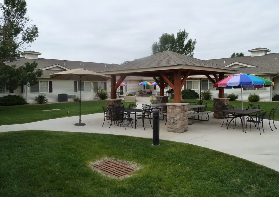 Outdoor patio area at a senior living facility with multiple tables and chairs under umbrellas and a wooden gazebo with stone pillars. The building surrounds the patio with windows and greenery around the area under a cloudy sky.