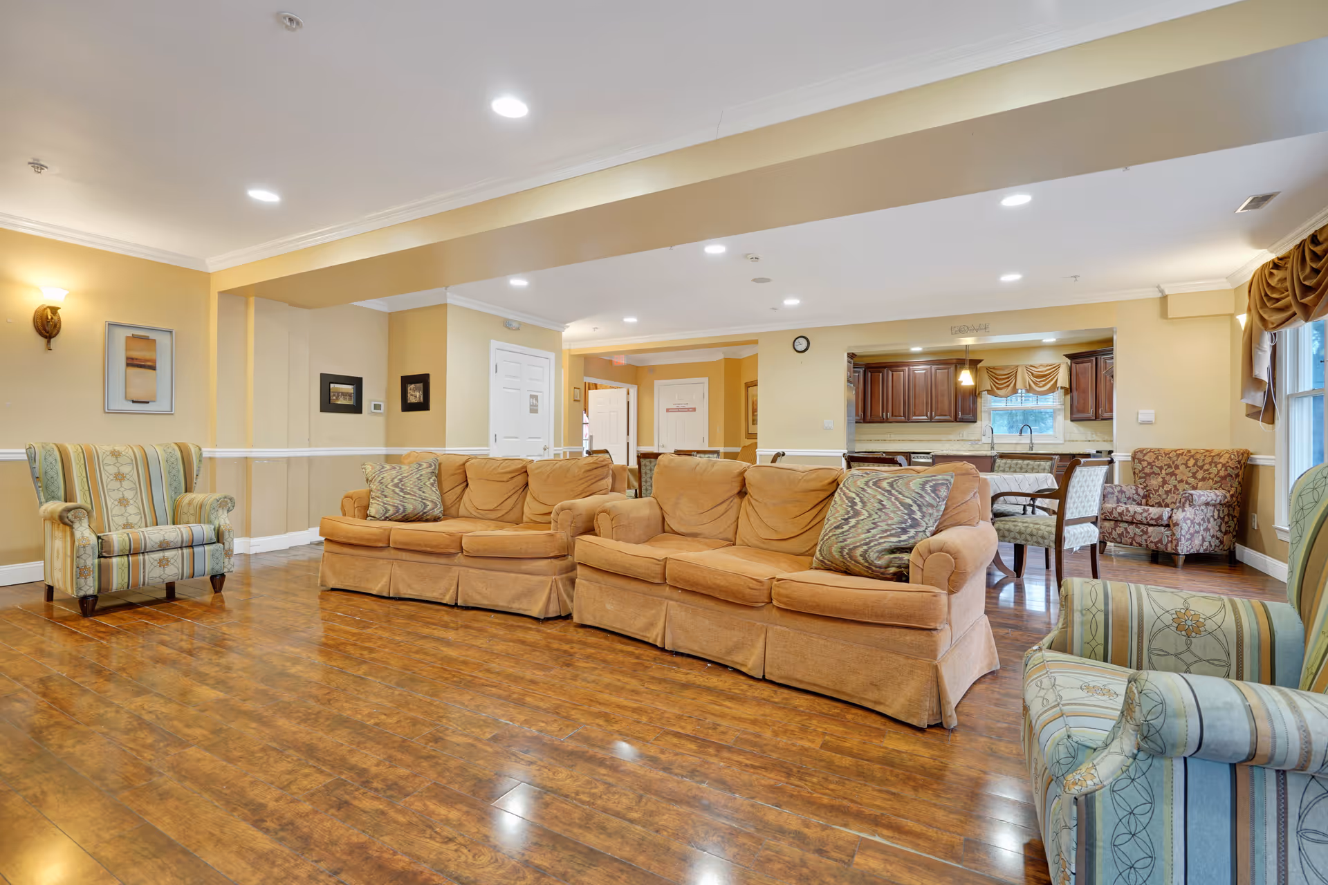 A spacious living room area in a senior living facility with wooden flooring, beige walls, and recessed lighting. The room features a large tan sofa with patterned cushions, two striped armchairs, and a floral armchair near a window with draped curtains. In the background, there is a dining area with a table and chairs, and a kitchen with wooden cabinets and a window above the sink.