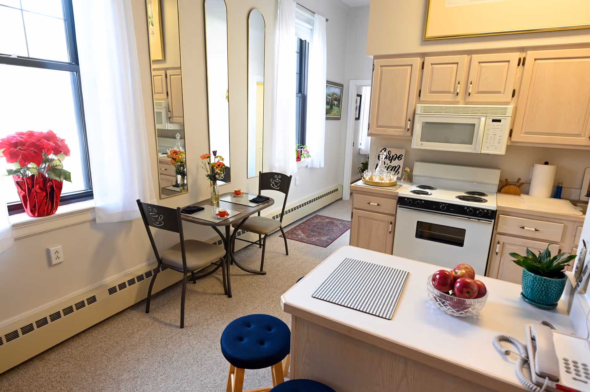 A bright kitchen and dining area with light wood cabinets, a white stove and microwave, a small table with two chairs, and a countertop with a bowl of apples and a potted plant. There are two tall mirrors on the wall behind the table, a window with white curtains, and a red flower pot on the windowsill.