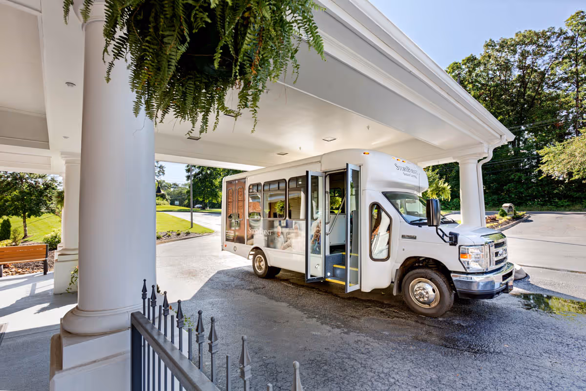 A white shuttle bus with the StoryPoint logo is parked under a covered entrance of a building, surrounded by greenery and trees on a sunny day.