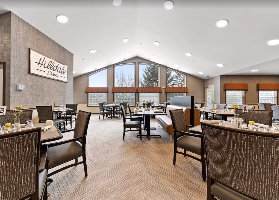 A spacious dining room with multiple tables and chairs arranged neatly. The room features large windows with brown valances allowing natural light to fill the space. A sign on the wall reads 'Hilldale Dining'. Tables are set with glasses, napkins, and small flower vases, creating a welcoming atmosphere.