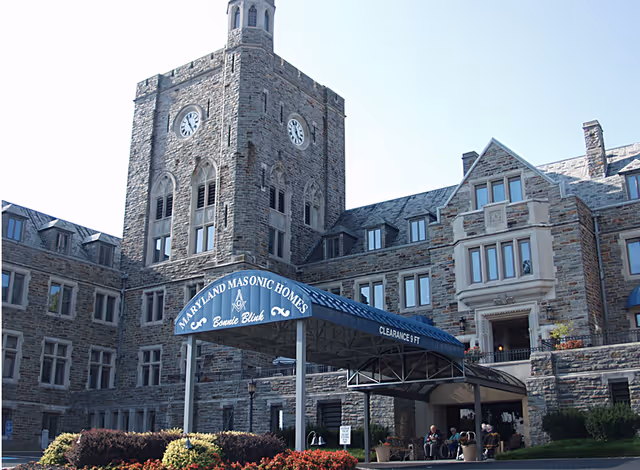 Stone building with a clock tower and multiple windows, featuring a blue canopy entrance labeled 'Maryland Masonic Homes Bonnie Blink' with people sitting near the entrance.