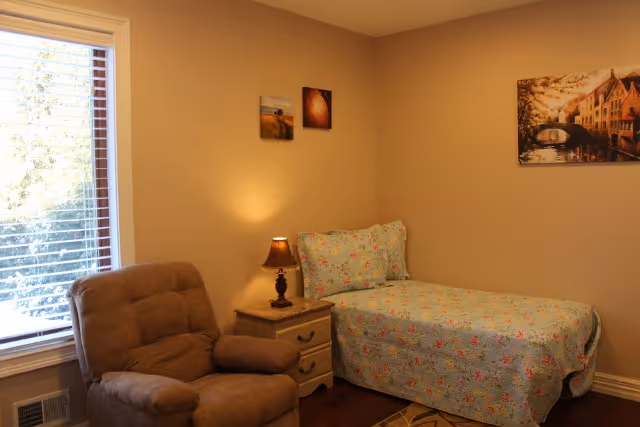 A cozy bedroom with a single bed covered in a floral quilt, a small wooden nightstand with a lamp, a comfortable brown recliner chair, and three framed pictures on the beige walls. A window with white blinds lets in natural light.