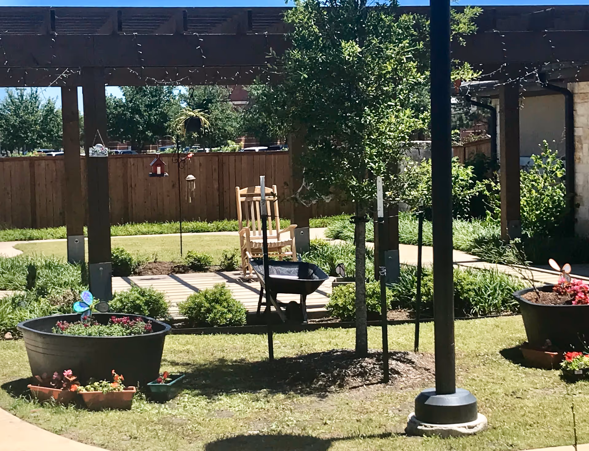 An outdoor garden courtyard with a wooden pergola, rocking chair, wheelbarrow and several large planters.