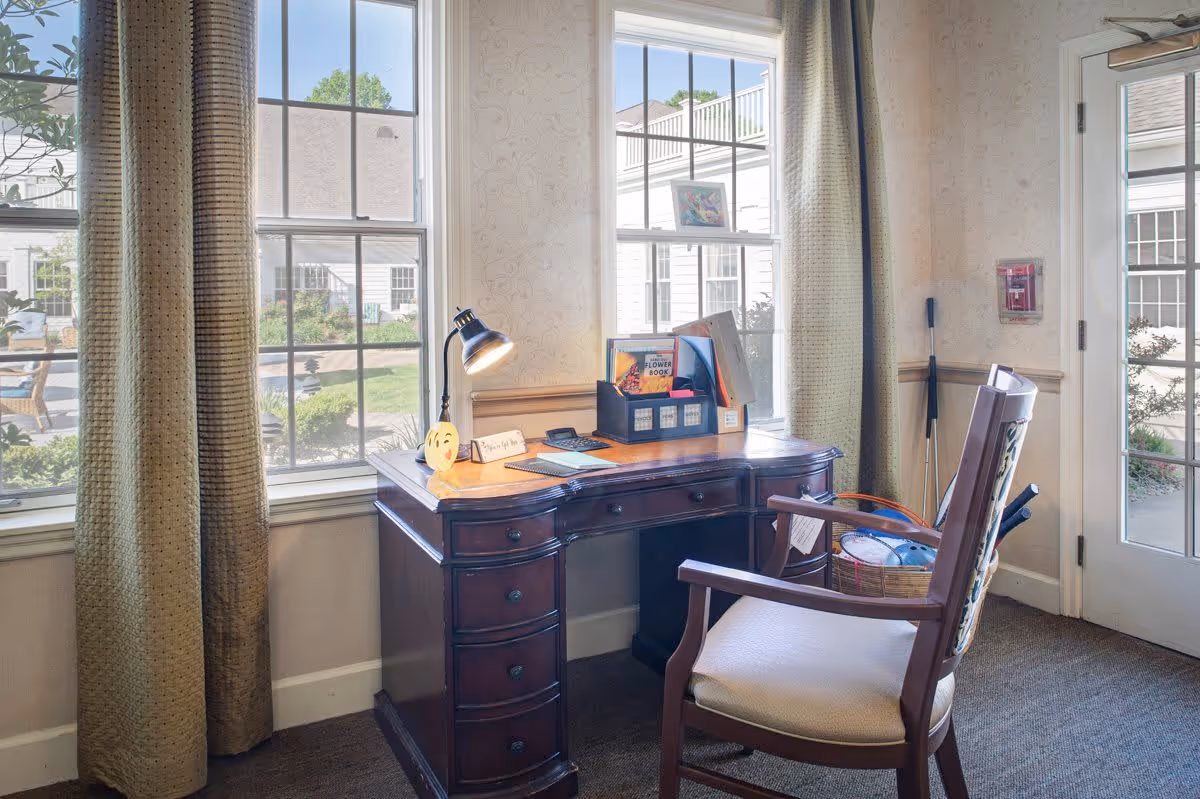 A cozy interior corner with a wooden desk and a cushioned wooden chair. The desk has a small lamp, a smiley face decoration, a calculator, and a black organizer holding books and papers. Two large windows with beige curtains let in natural light, showing an outdoor garden area. A door with glass panels is visible on the right side.
