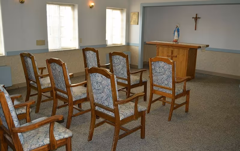 A small chapel or prayer room with wooden chairs arranged in rows facing a wooden altar. On the altar is a statue of the Virgin Mary and a cloth. A crucifix is mounted on the wall above the altar. The room has two windows with blinds and soft lighting from wall sconces.