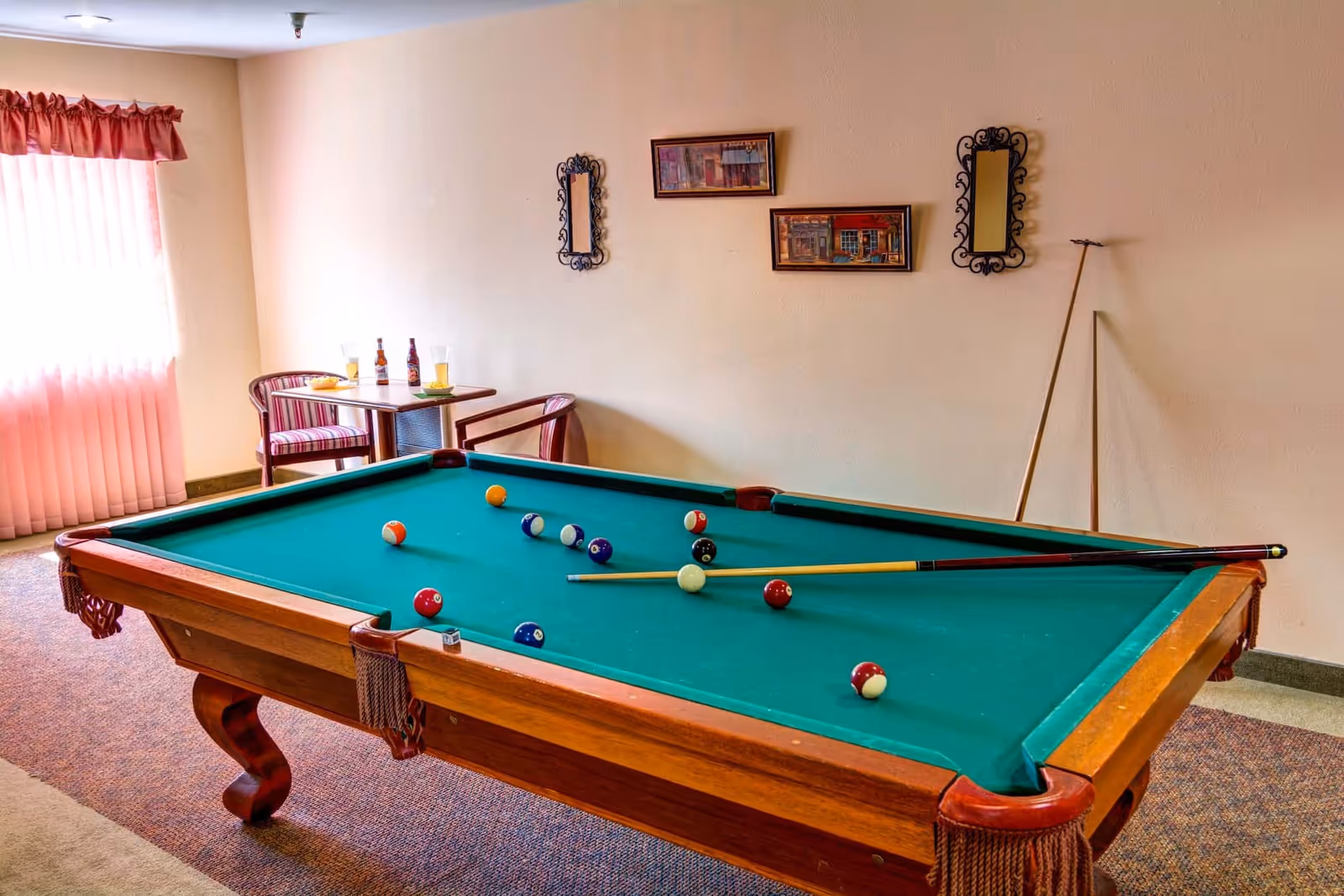 A billiards table with cues and scattered balls in a well-lit recreation room with a small table and chairs by a curtained window.