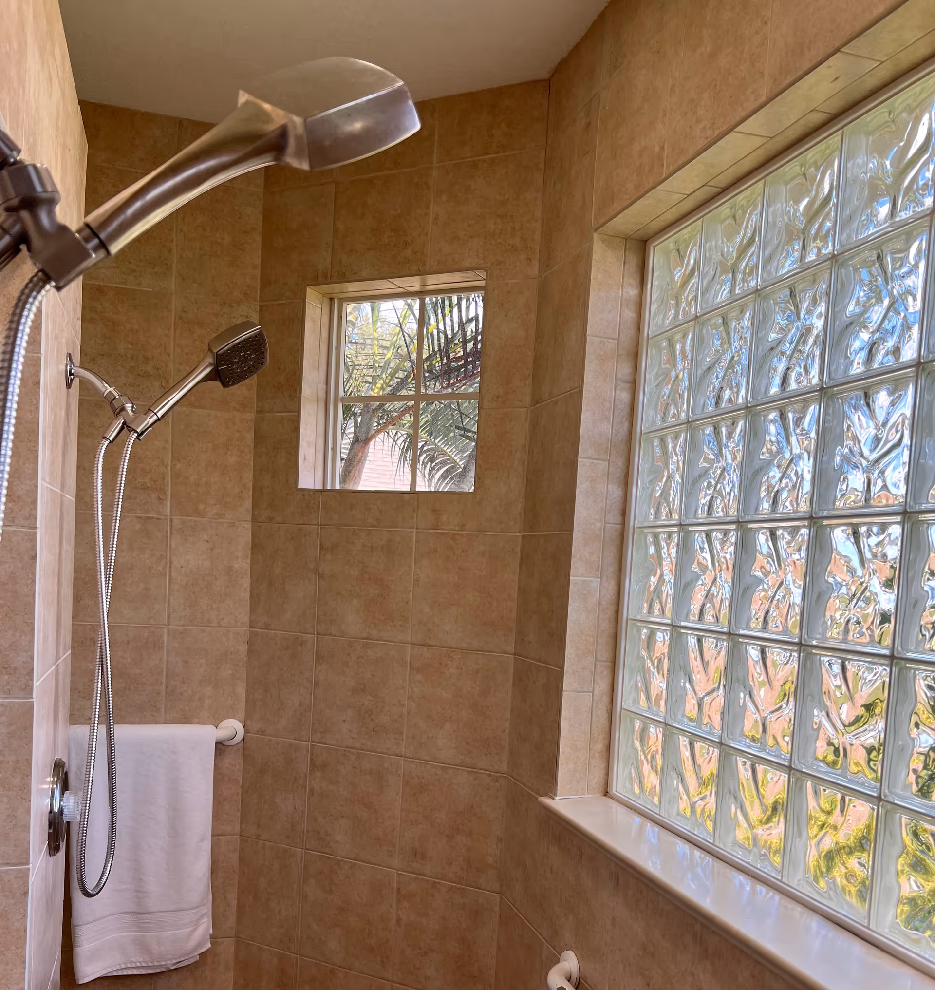Tiled walk-in shower with dual handheld showerheads, a towel on a bar, and a large glass-block window letting in light.