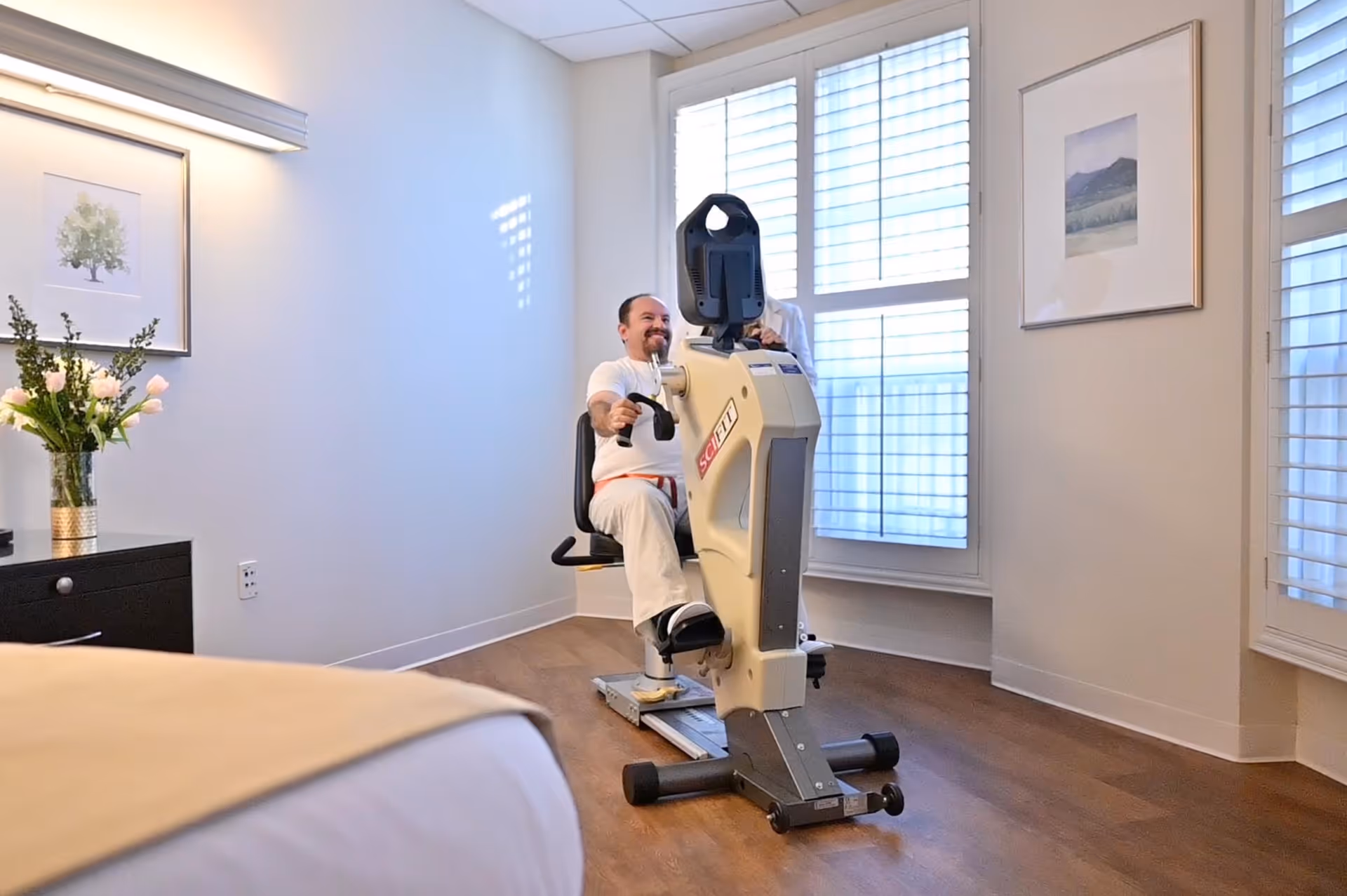 A man is seated on a recumbent exercise machine in a bright room with wooden floors and large windows with white blinds. The room has light-colored walls, framed artwork, a black dresser with a vase of flowers, and a bed partially visible in the foreground.