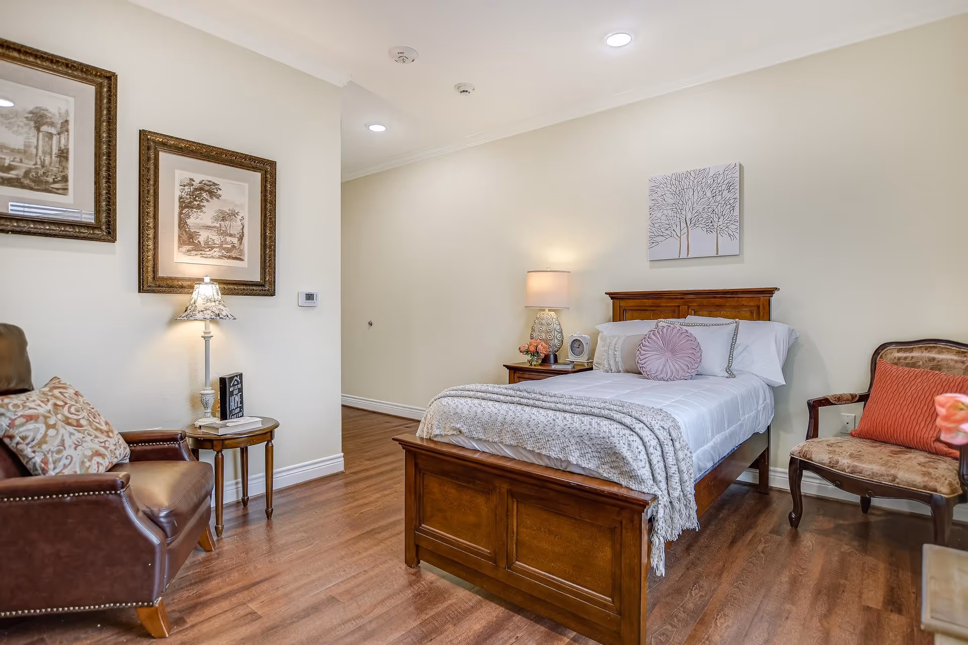 A cozy bedroom in La Fontaine Memory Care featuring a wooden bed with white bedding and decorative pillows, a wooden nightstand with a lamp and flowers, a leather armchair with a patterned cushion, a small round side table with a lamp and books, framed artwork on the wall, and hardwood flooring.