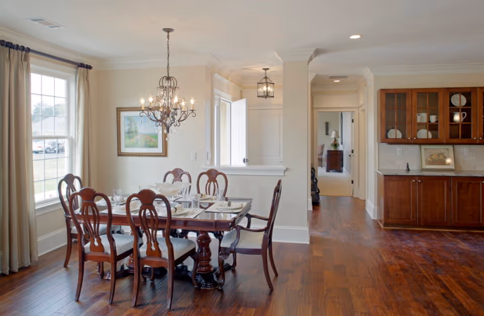 A bright dining room with a wooden dining table set for six, featuring elegant chairs with cushioned seats. A decorative chandelier hangs above the table. The room has large windows with beige curtains, wooden flooring, and a built-in wooden cabinet with glass doors displaying dishes. The space opens into a hallway leading to other rooms.