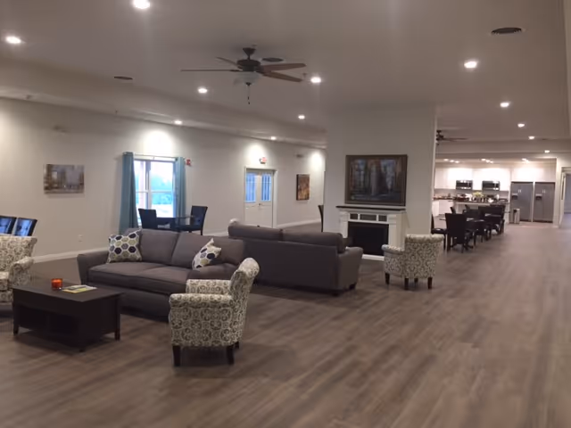 Spacious interior of a senior living facility featuring a large open area with multiple seating arrangements including sofas and patterned armchairs. There is a fireplace with a framed painting above it, and in the background, a dining area with tables and chairs is visible next to a kitchen equipped with stainless steel appliances. The room has wood flooring, recessed lighting, and ceiling fans.